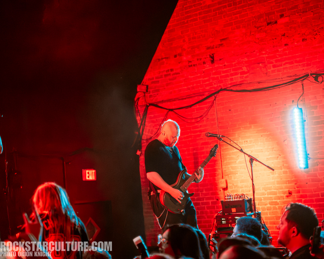 A guitarist performing on stage with dramatic red lighting, while an audience watches in the foreground.