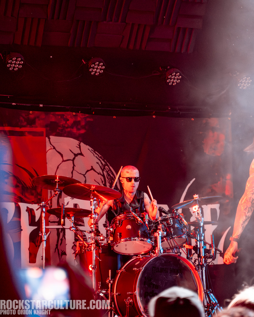A drummer performing on stage, wearing sunglasses and a sleeveless vest, surrounded by smoke and colorful stage lighting.