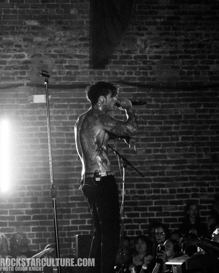 A performer with tattoos stands on stage, holding a microphone while singing. The background features exposed brick walls, and an audience watches intently.