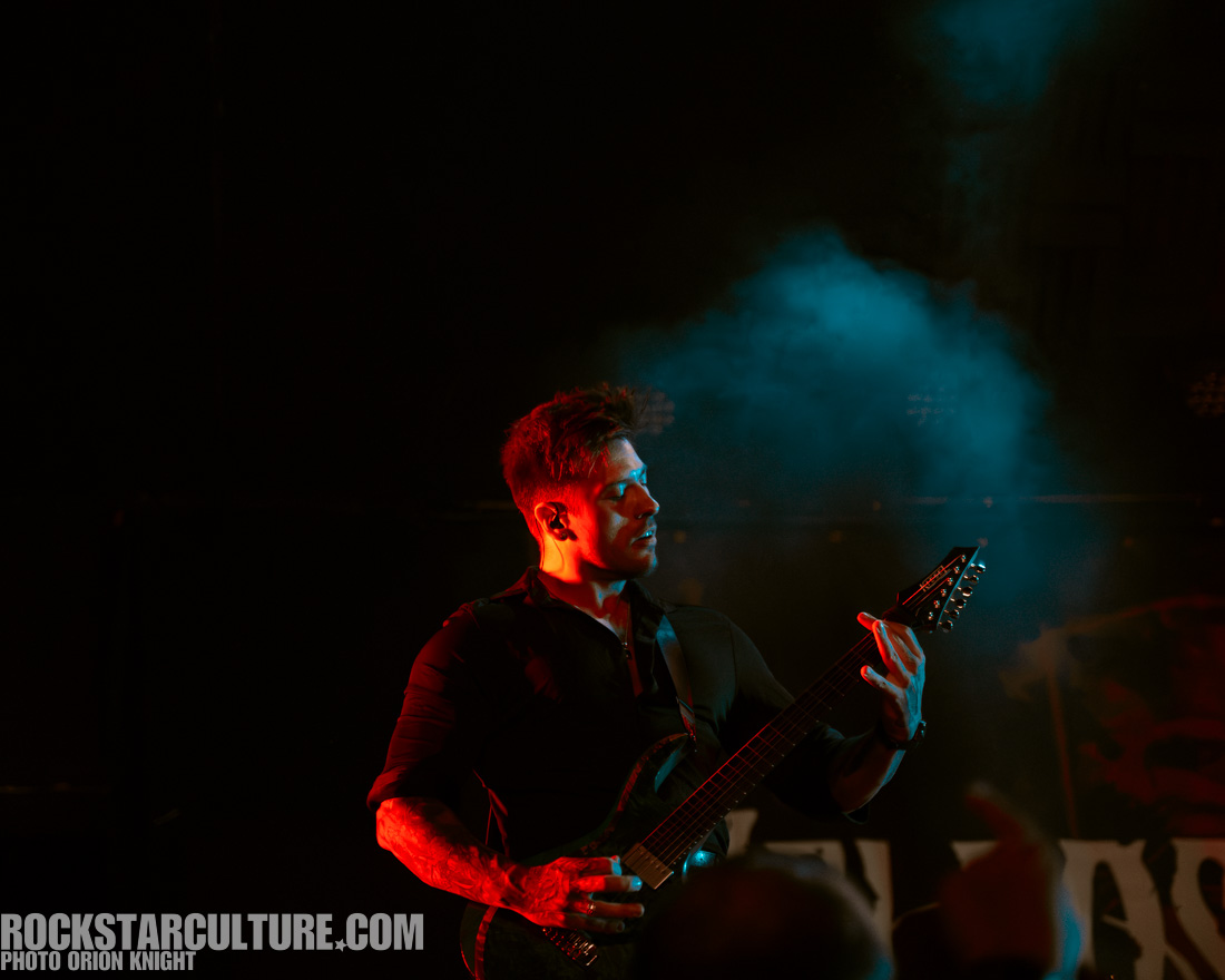 A musician playing an electric guitar on stage, illuminated by colorful lighting with smoke in the background.