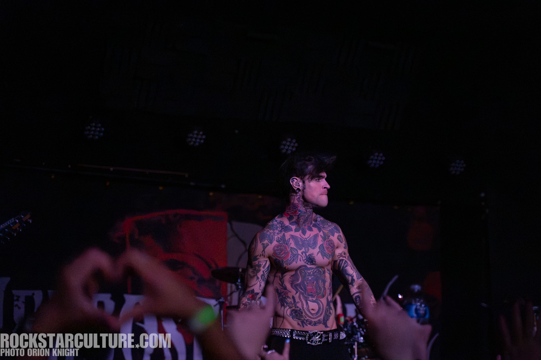 A tattooed male performer on stage, displaying various tattoos on his torso and arms, with a close-up view of the audience making a heart shape with their hands in the foreground.