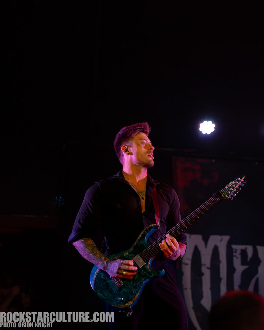 A musician passionately playing an electric guitar on stage, wearing a black shirt and with colorful stage lighting in the background.