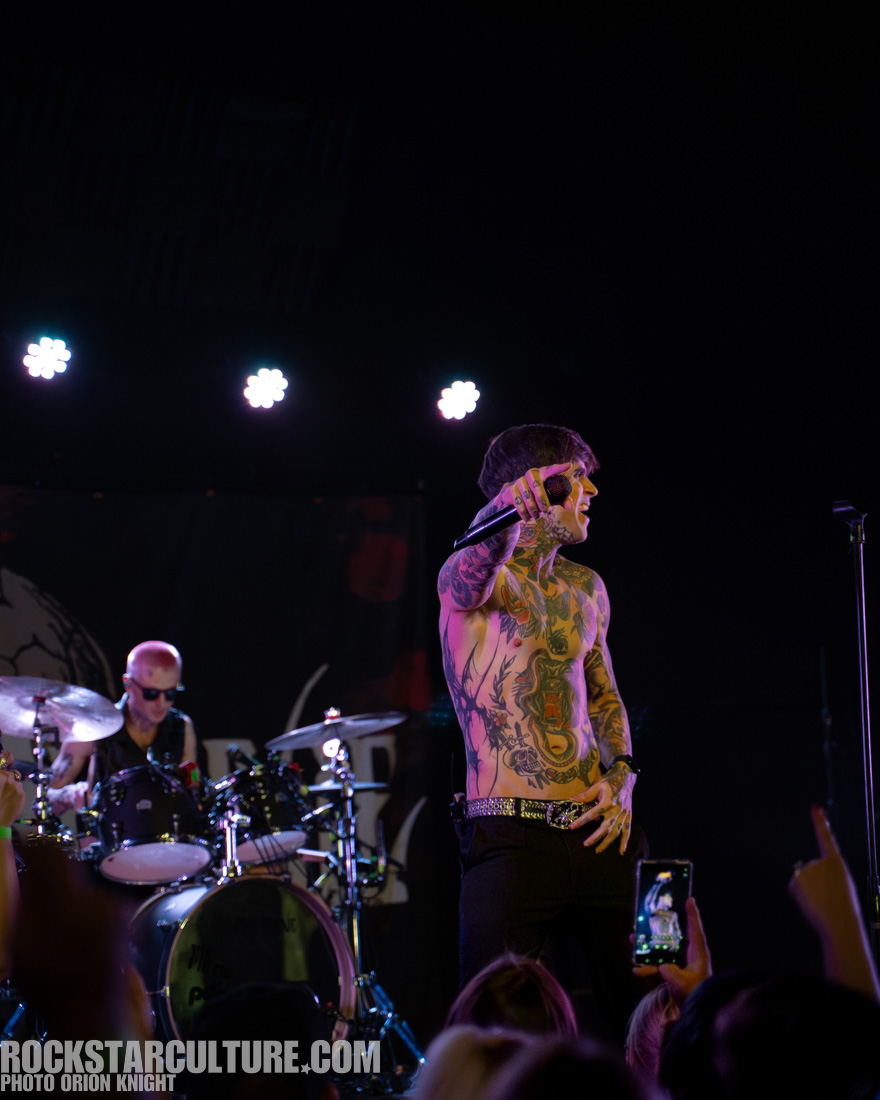 A shirtless singer with tattoos performs on stage, holding a microphone and gesturing to the audience, while a drummer plays in the background.