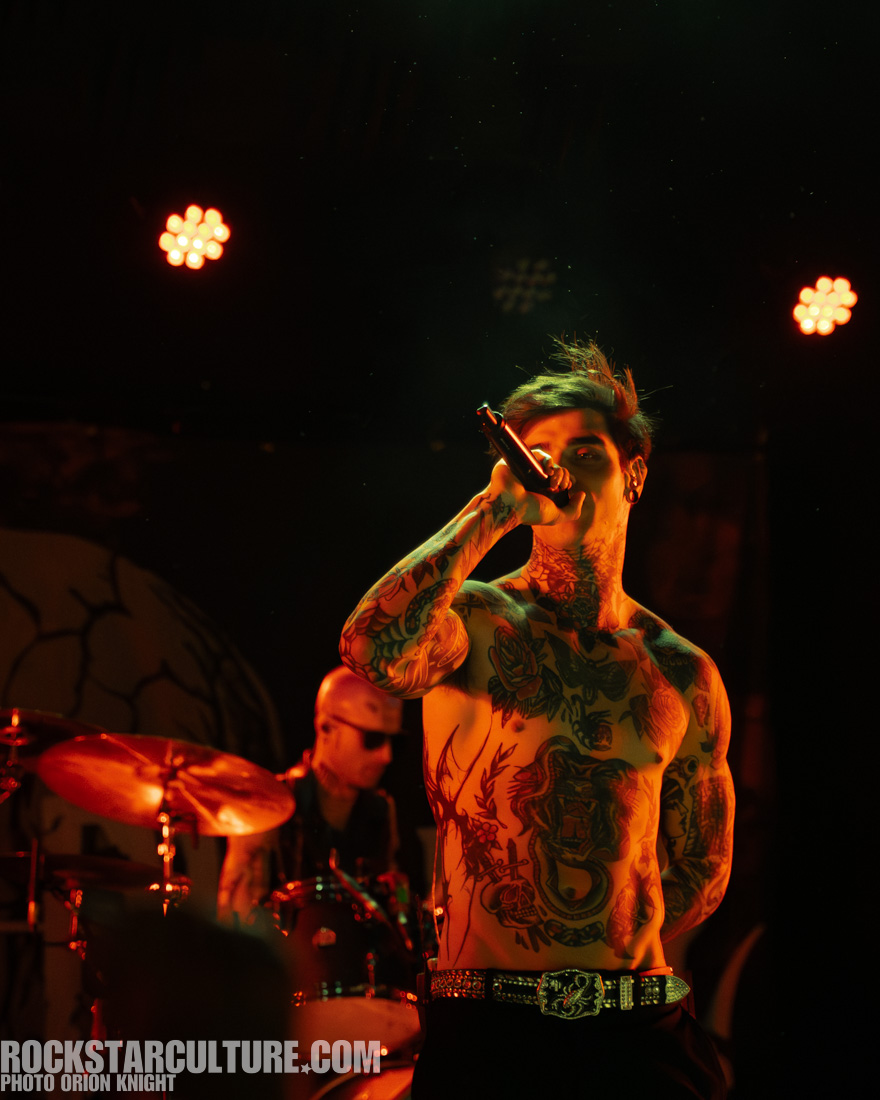 Male vocalist with tattoos performing on stage, holding a microphone, with a drummer in the background.