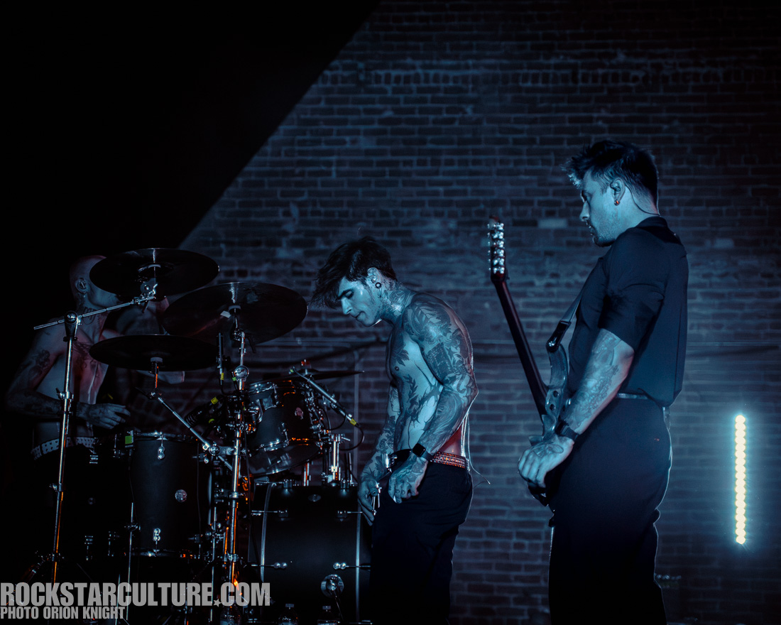 A rock band performing on stage, featuring a drummer and two guitarists. The musicians have tattoos and are engrossed in their performance. The lighting is dim with blue tones, highlighting their intense expressions.