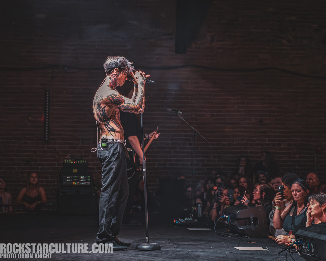 A tattooed male singer performing on stage with a microphone, while an audience watches intently.