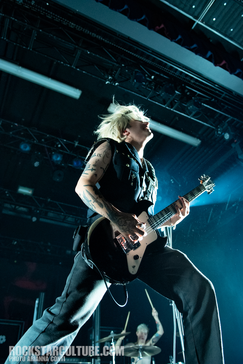 A musician with short blonde hair passionately playing an electric guitar on stage, captured in mid-performance with bright stage lights and a drummer visible in the background.