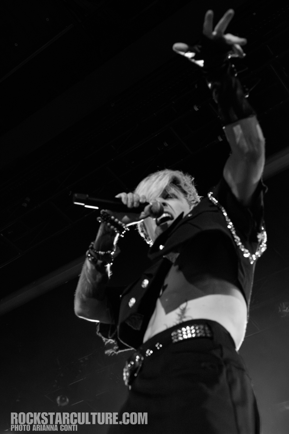 A performer on stage passionately singing into a microphone, wearing a cropped jacket and adorned with jewelry, captured in black and white.