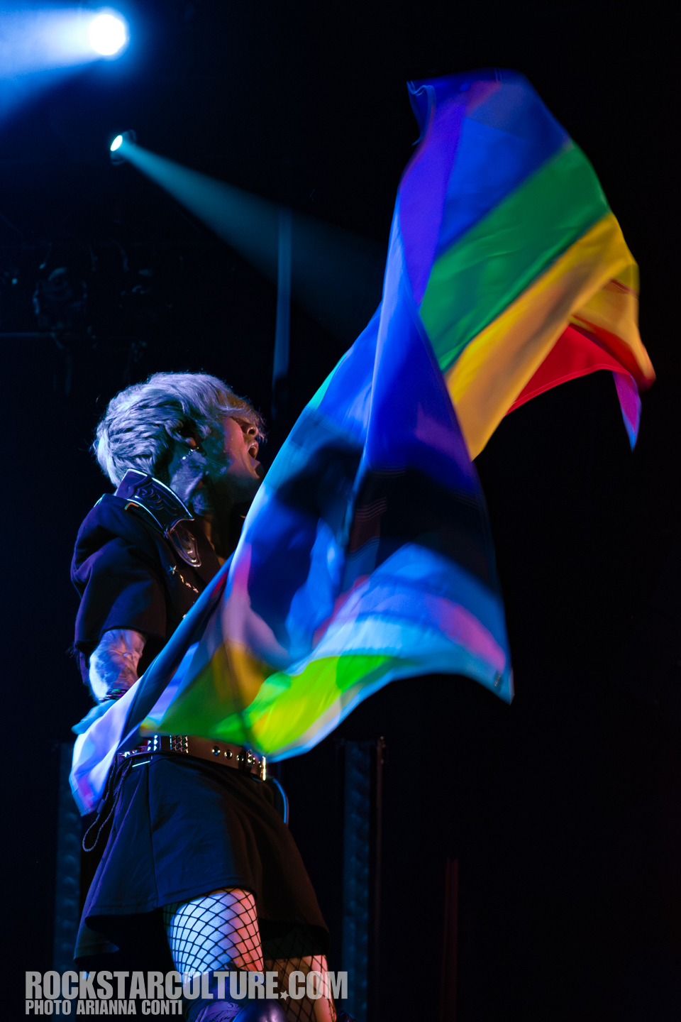 A performer passionately waving a colorful pride flag on stage, wearing a black outfit with fishnet stockings and a dynamic hairstyle, illuminated by stage lights.