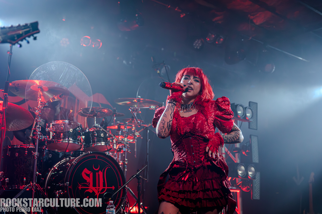 A female singer with red hair and a red dress performs on stage, holding a microphone, with a drum set and stage lights in the background.