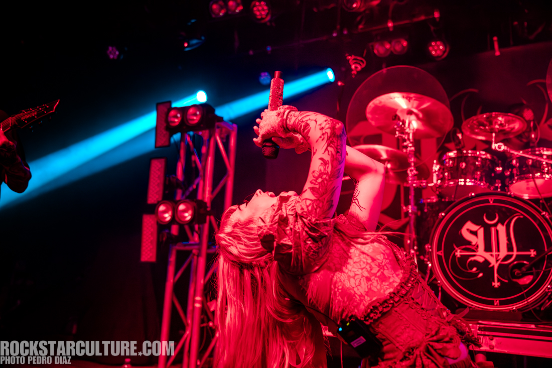 A performer passionately singing into a microphone with elaborate stage lighting and a drum set in the background.