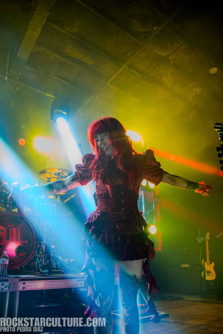 A performer with red hair in a striking outfit stands on stage, surrounded by colorful stage lights and smoke.
