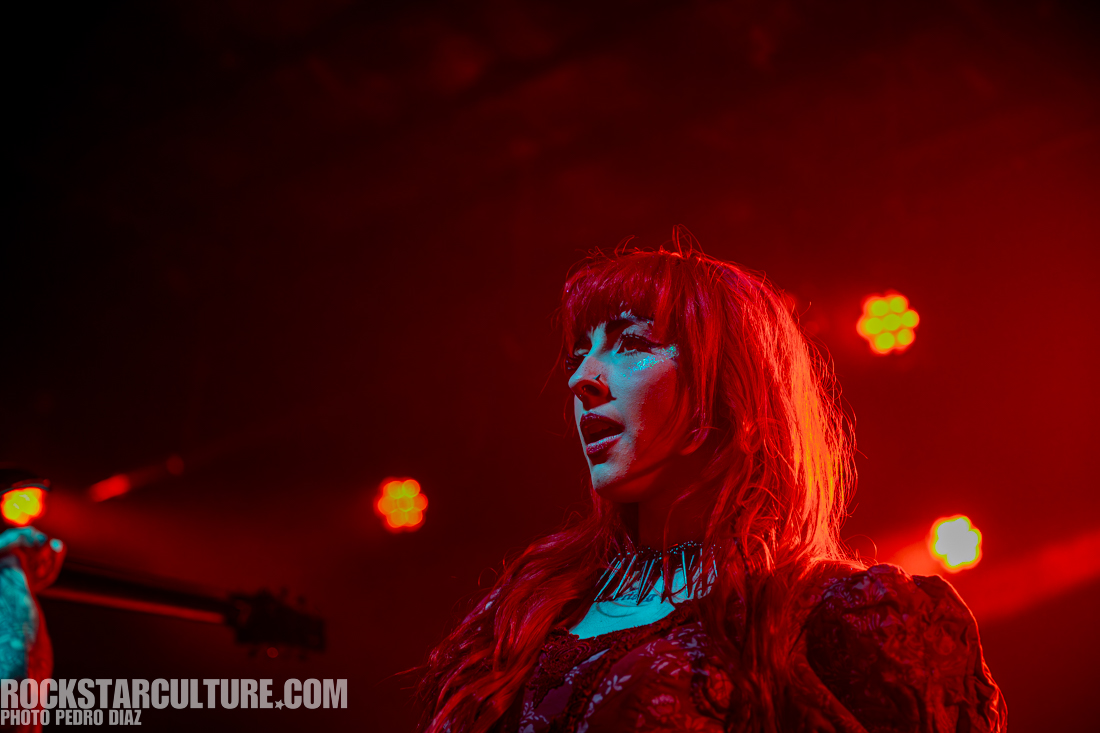 Close-up of a woman with bright red hair, wearing dark clothing and a choker, performing on stage under red lighting.