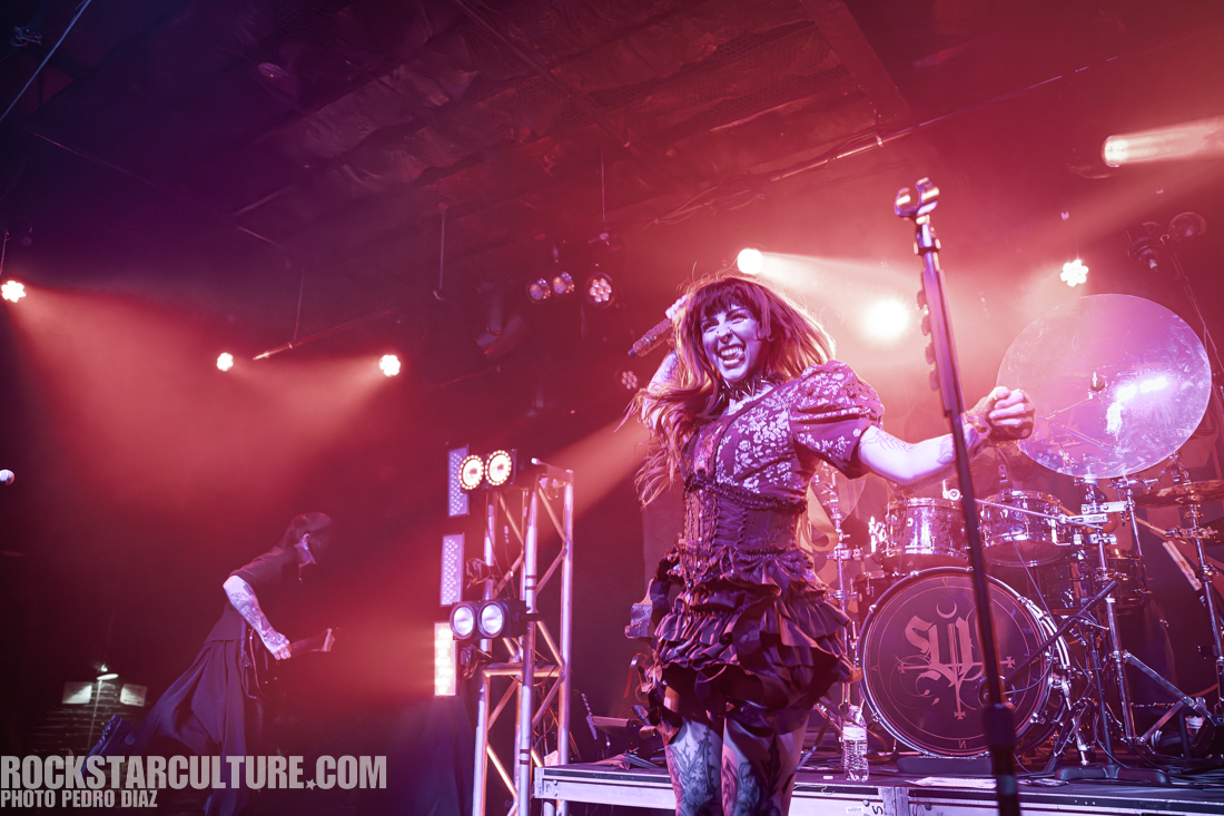 A female performer on stage, wearing a black and purple dress with a lively expression, singing into a microphone while illuminated by stage lights. A band member is visible in the background with instruments set up behind them.