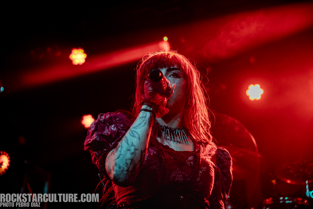 A close-up of a female singer with red hair and tattoos, holding a microphone and performing on stage with dramatic lighting.