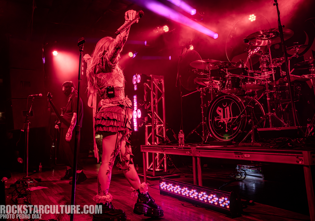 A singer with long hair and tattoos performs energetically on stage, holding a microphone aloft, while a guitarist and drummer are in the background. The stage is illuminated with vibrant pink and purple lights.