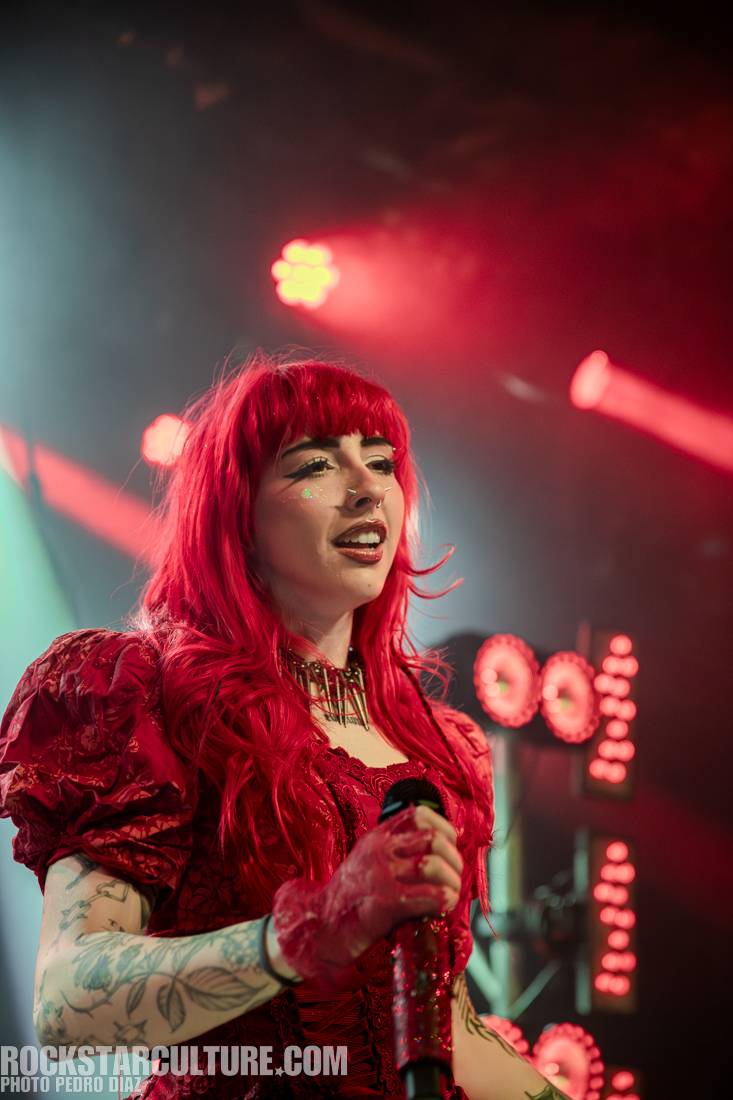 A female performer with bright red hair sings passionately on stage, wearing a red dress with puffed sleeves and holding a microphone. She has tattoos visible on her arms and sparkling makeup on her face, surrounded by colorful stage lights.