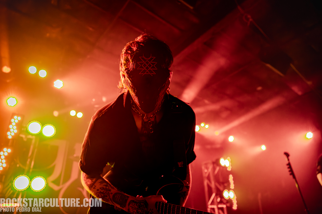 A musician performing on stage, wearing a mask and a dark shirt, playing an electric guitar under dramatic lighting with red and green hues.