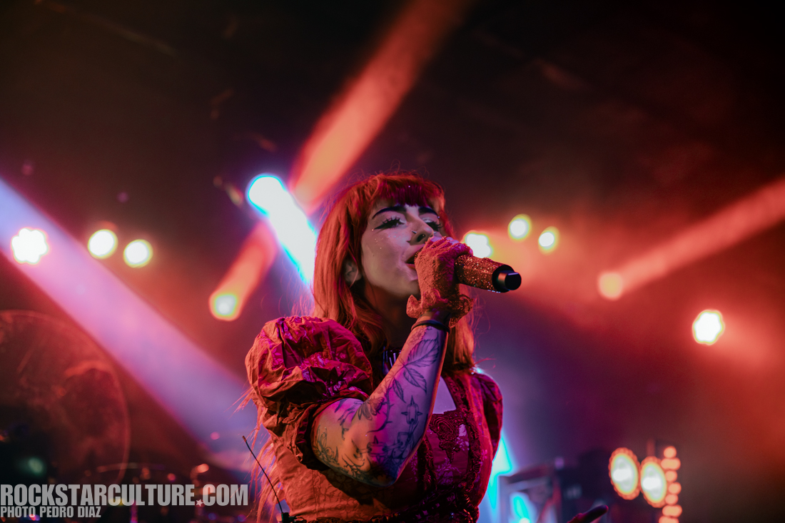A female singer with red hair performs on stage, wearing a glittery red outfit and holding a microphone surrounded by colorful stage lights.