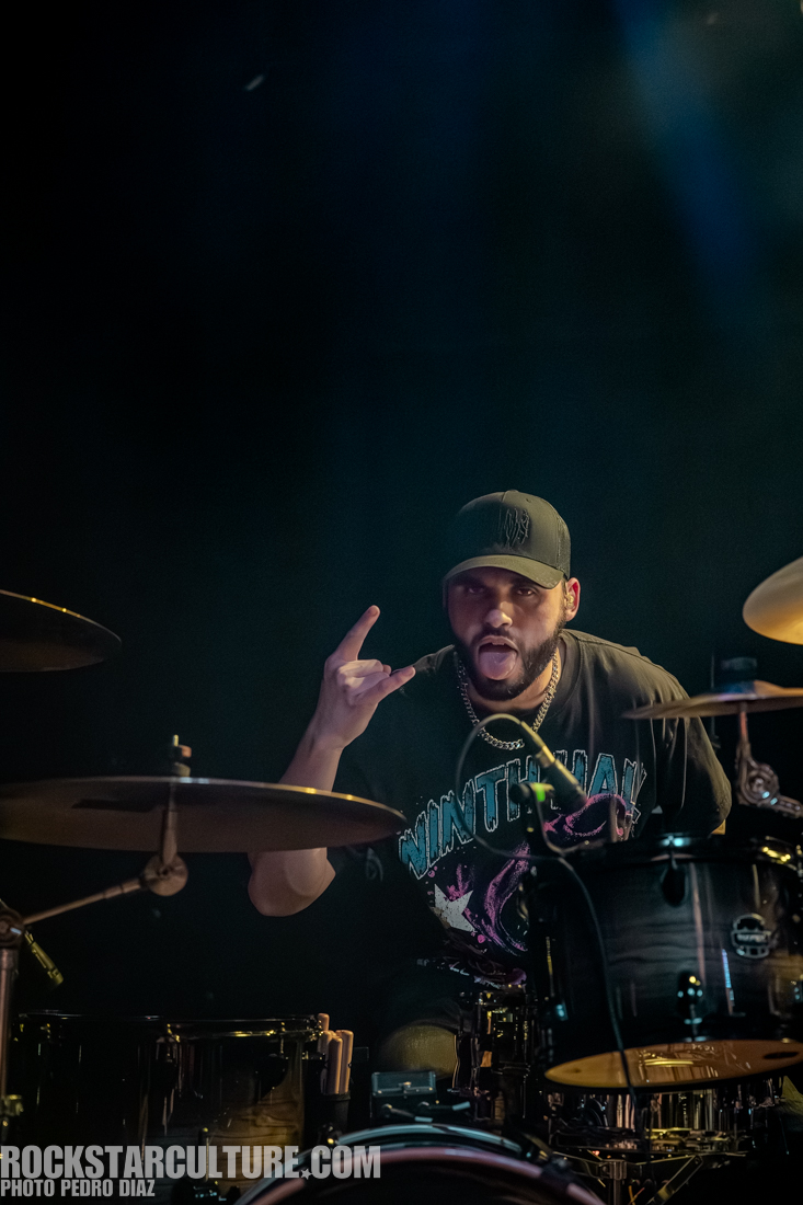 A drum player performing energetically on stage, making a rock hand gesture and sticking out his tongue.