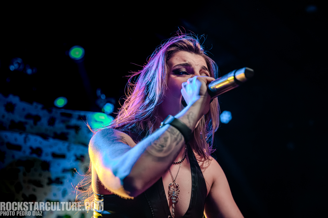 A performer singing into a microphone on stage, with colorful lights in the background.
