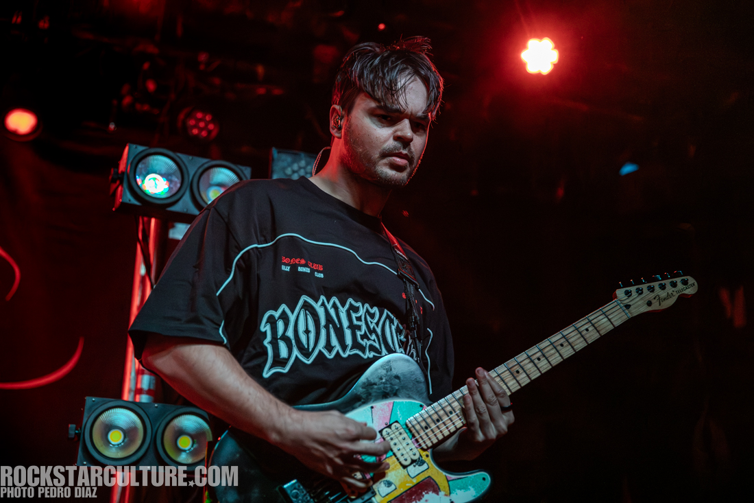 A musician playing a colorful electric guitar on stage, wearing a black 'BONES' t-shirt, with stage lighting illuminating the scene.