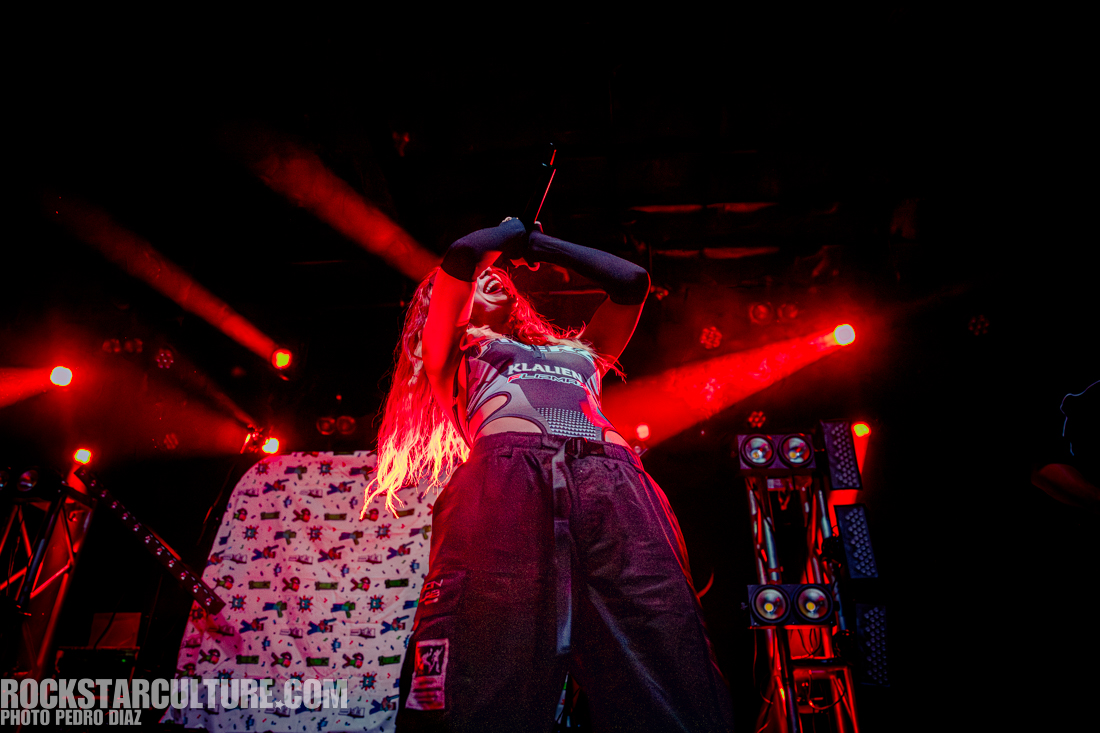 A female performer on stage with long, flowing hair, wearing a stylish outfit, passionately singing into a microphone under dramatic red stage lights.