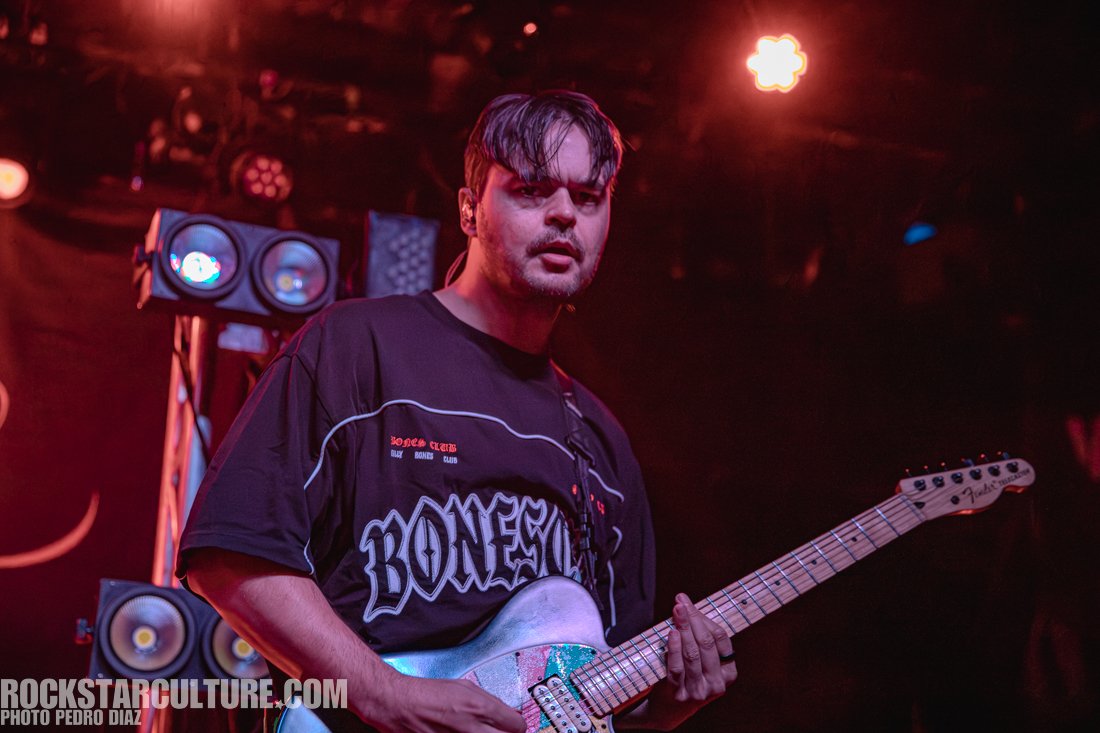 A musician performing on stage holding an electric guitar, wearing a black graphic t-shirt, with stage lights illuminating the background.
