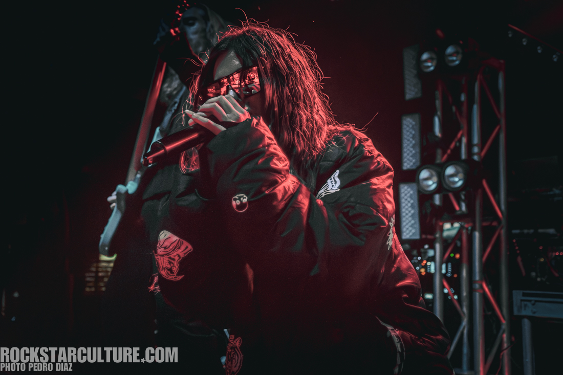 A singer wearing sunglasses and a black oversized jacket passionately performs on stage with a microphone, surrounded by stage lights and a dimly lit background.