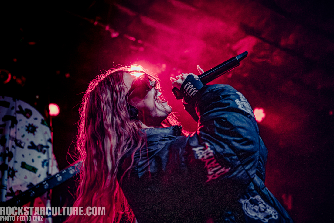 A performer singing passionately on stage, wearing sunglasses and a dark jacket, surrounded by dramatic pink lighting.