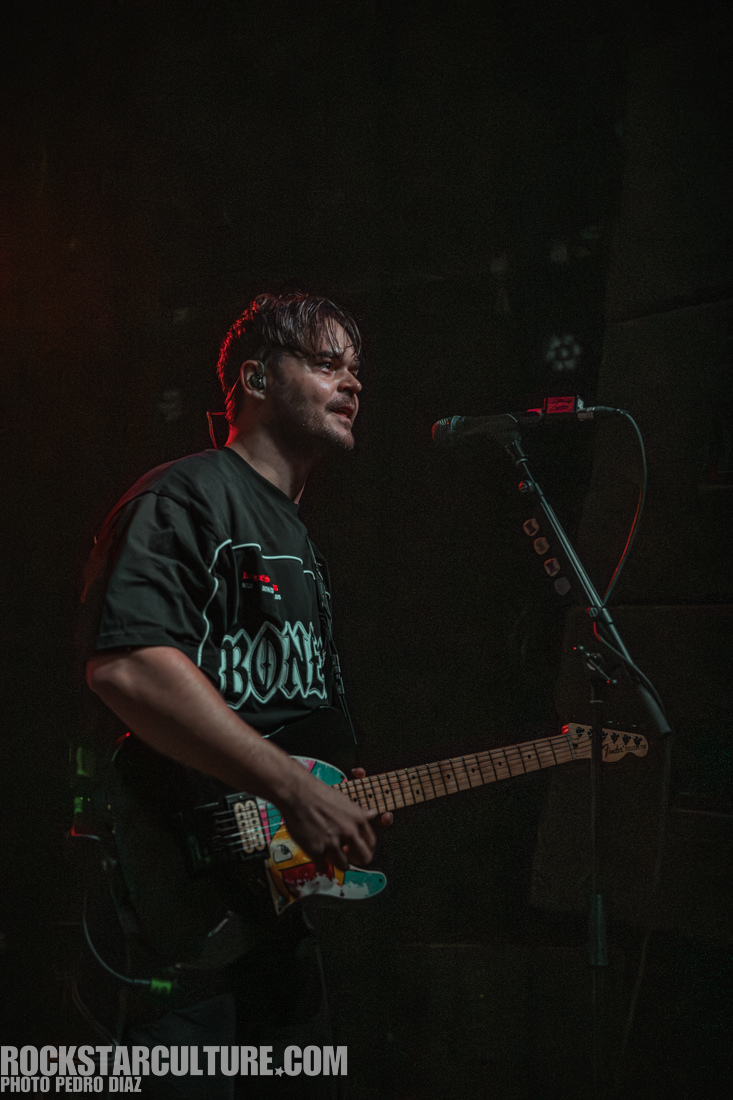 A musician performing on stage, playing an electric guitar, wearing a black graphic t-shirt with the word 'BONE' and standing next to a microphone.