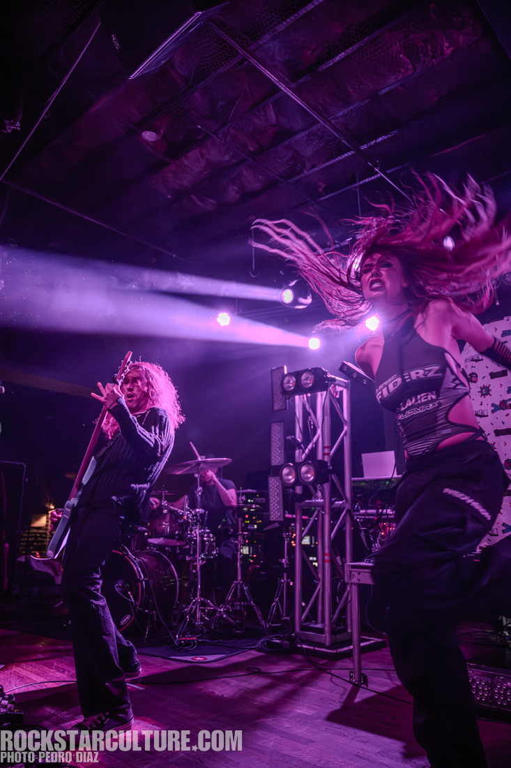 A dynamic live performance featuring a guitarist and a vocalist energetically engaging with the audience, illuminated by vibrant stage lights.