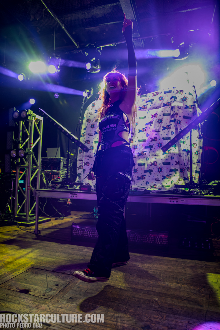A performer on stage with vibrant lighting, smiling and pointing upwards, wearing stylish stage attire, with a backdrop featuring colorful graphics.