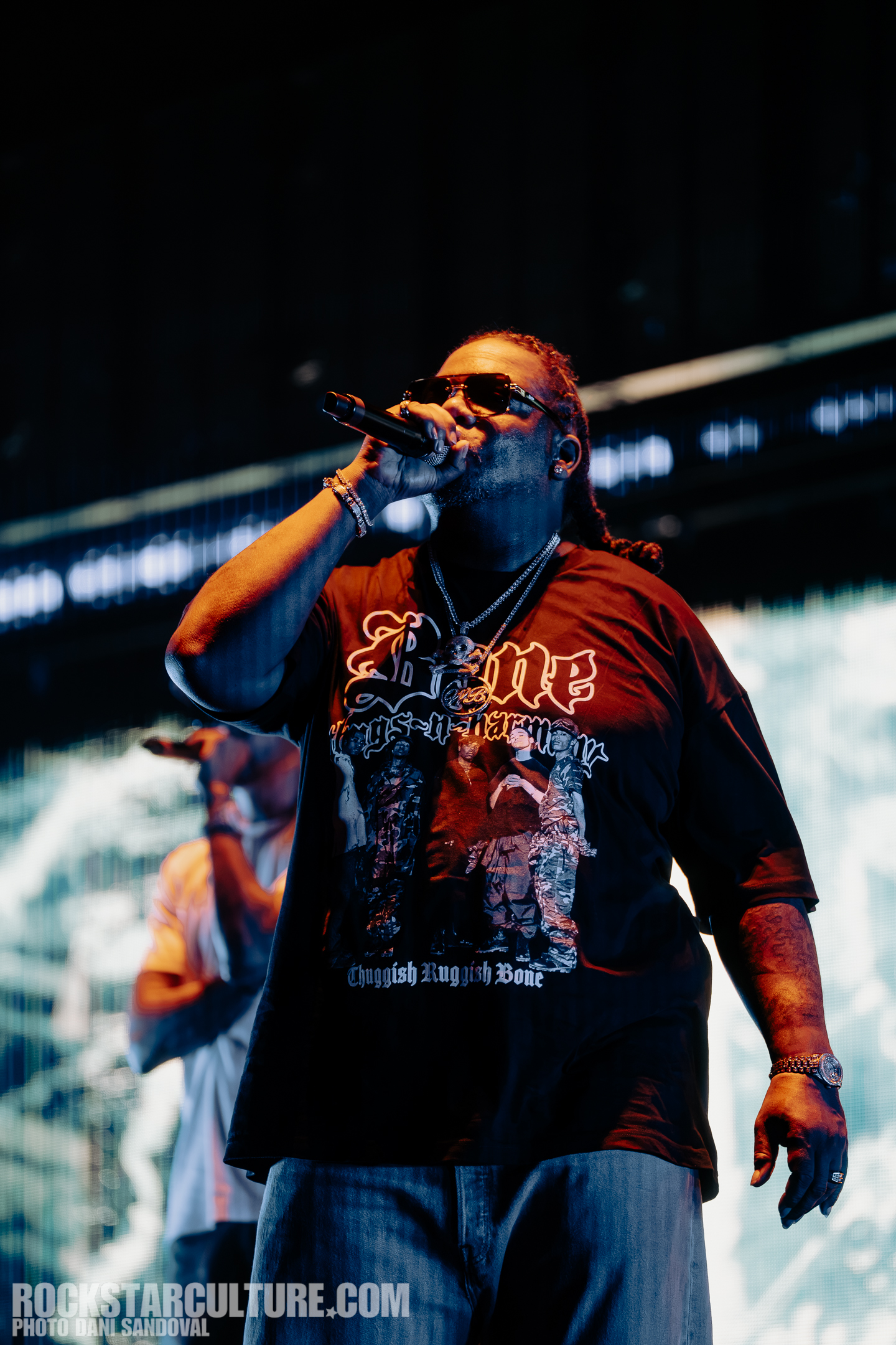 A male vocalist performing on stage, wearing sunglasses and a graphic t-shirt with a prominent logo. He is holding a microphone and is in the foreground of a blurred background, indicating a lively concert atmosphere.
