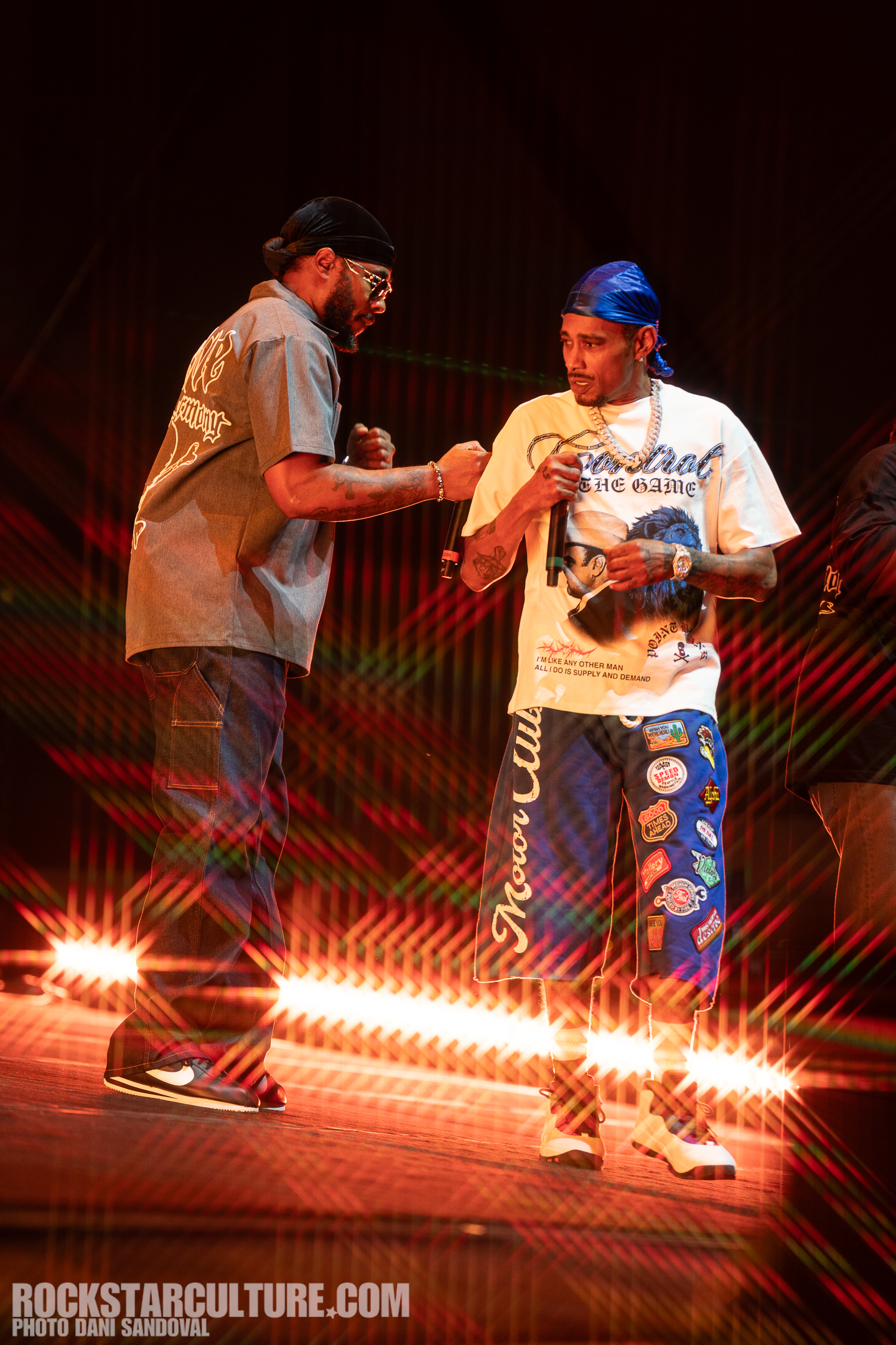 Two men performing on stage, one wearing a gray shirt and sunglasses, the other in a white graphic T-shirt and blue pants with patches, both holding microphones.