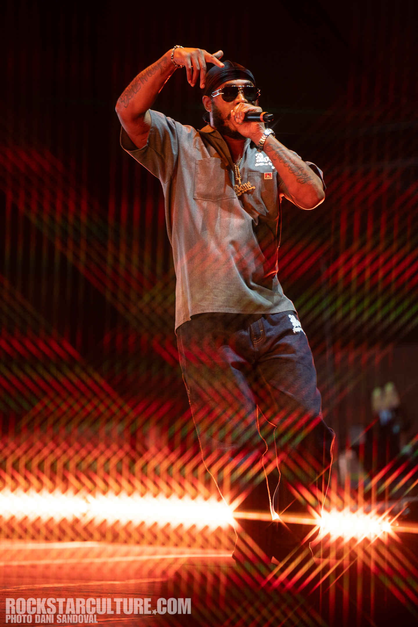 A performer on stage wearing sunglasses and a bandana, gesturing while holding a microphone, with colorful laser lights and vibrant stage effects in the background.