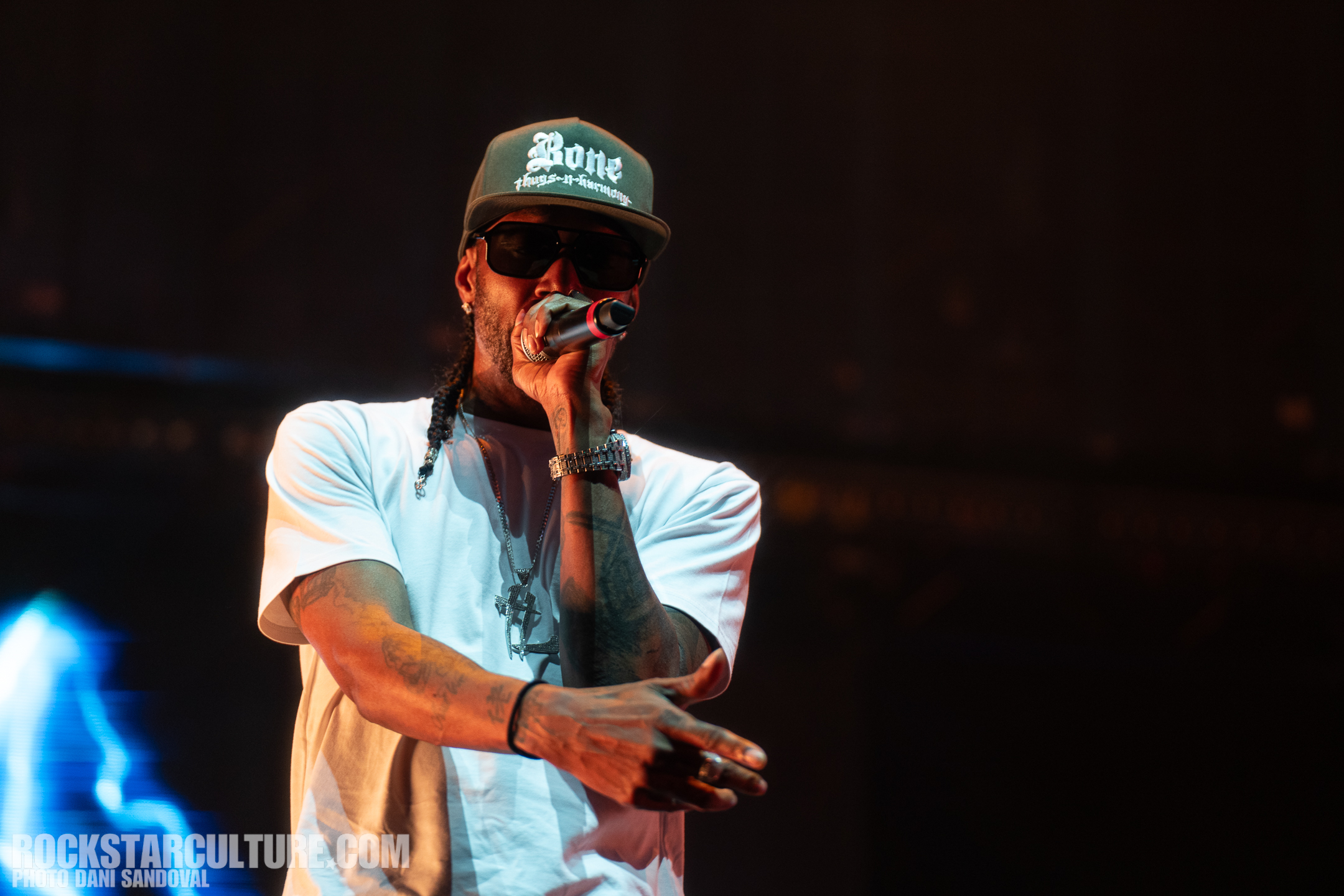A musician performing on stage, wearing a white shirt and a green cap, holding a microphone and gesturing with his hands.