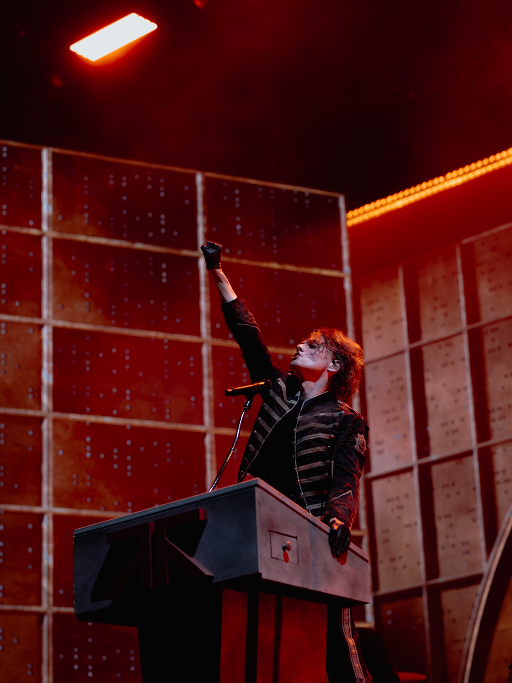 A performer with dramatic makeup and a military-style jacket stands at a microphone, raising one arm in a powerful pose on stage, with a backdrop of illuminated panels and a red lighting atmosphere.