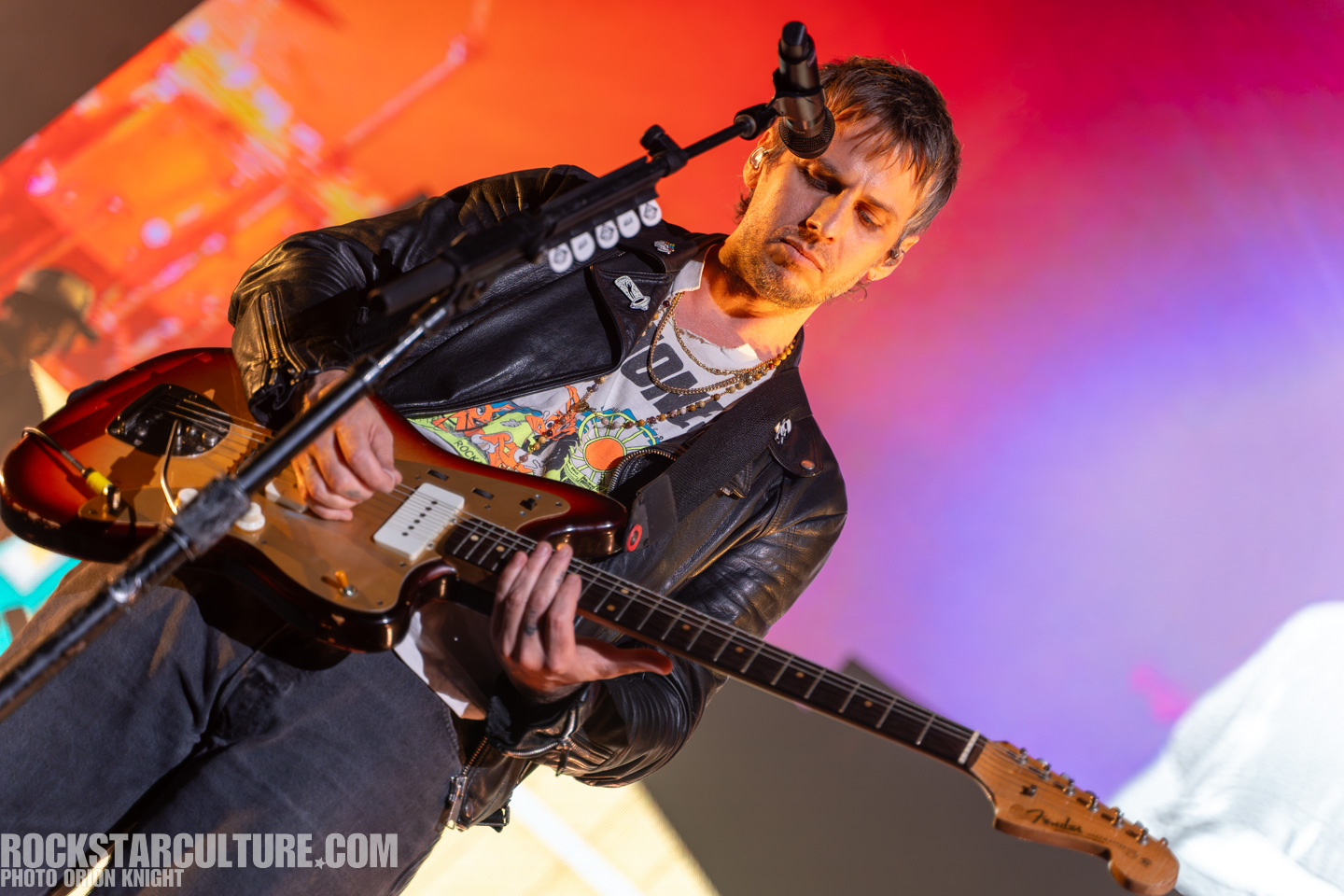 Musician playing an electric guitar on stage, wearing a leather jacket and colorful t-shirt, with vibrant stage lighting in the background.