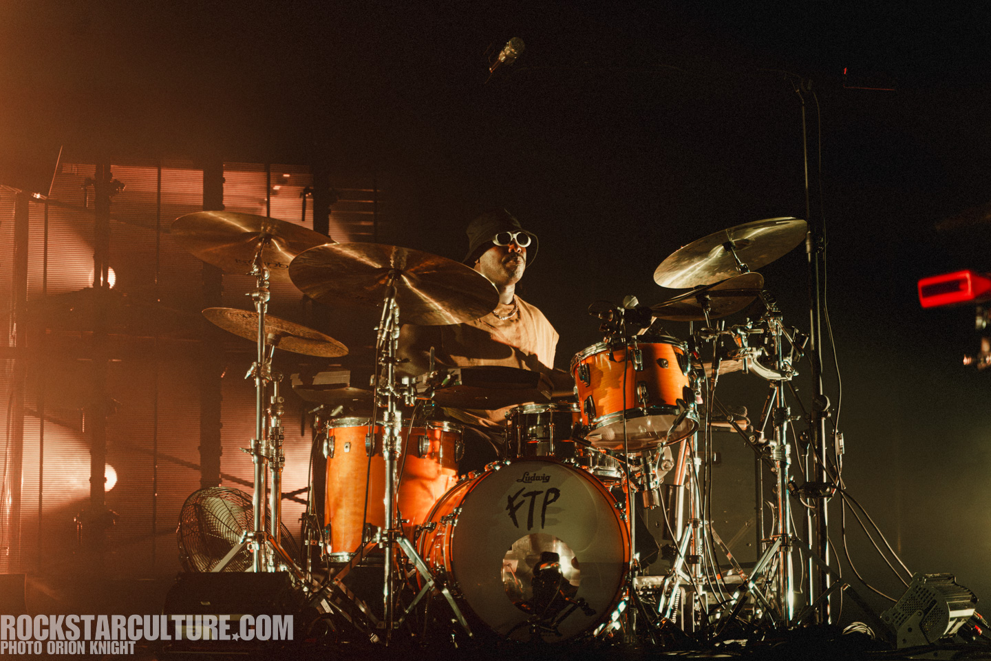 Drummer performing on stage with an orange drum set, wearing sunglasses and a hat, with dramatic lighting in the background.