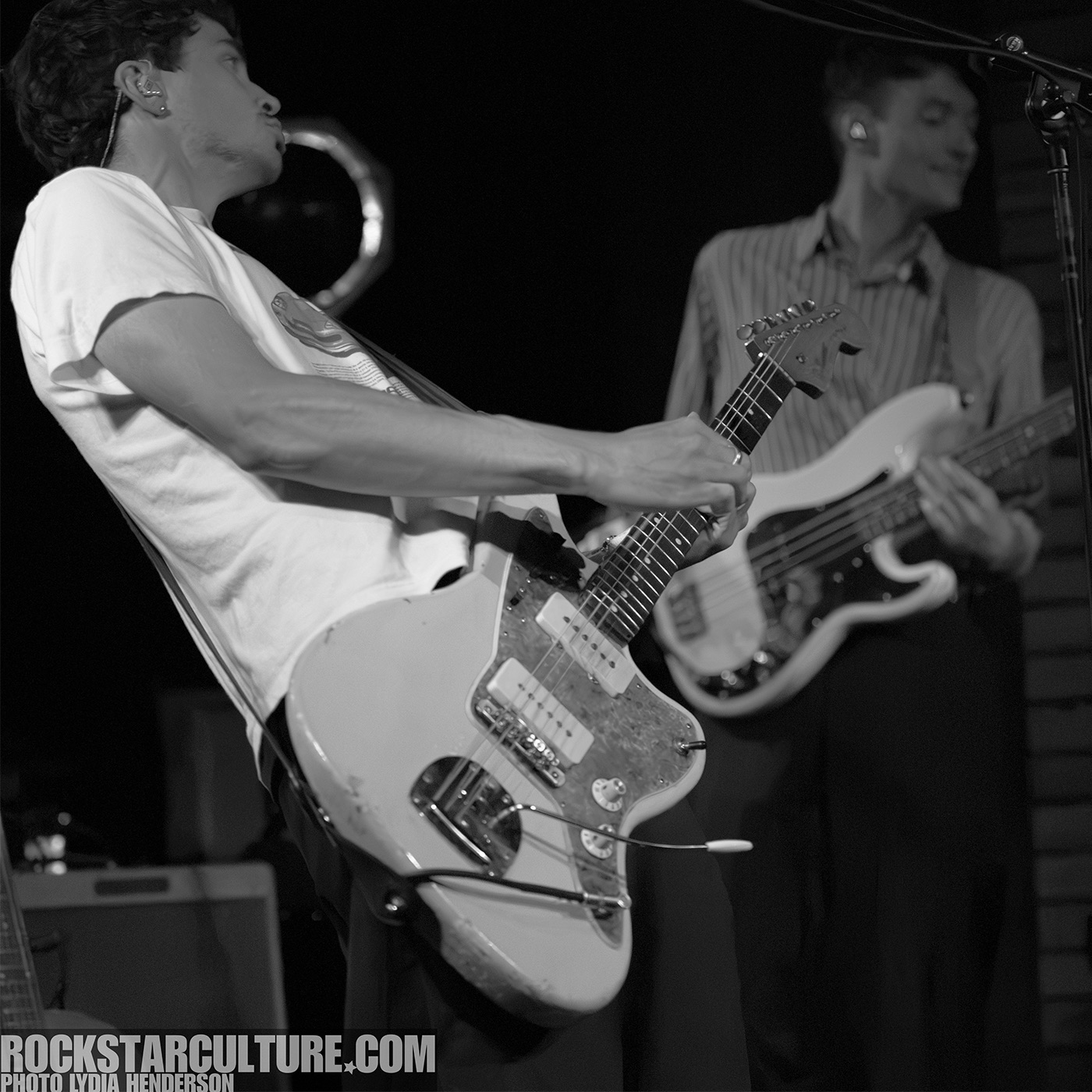 A guitarist passionately playing a light-colored electric guitar on stage, with another musician playing bass in the background, both captured in black and white.