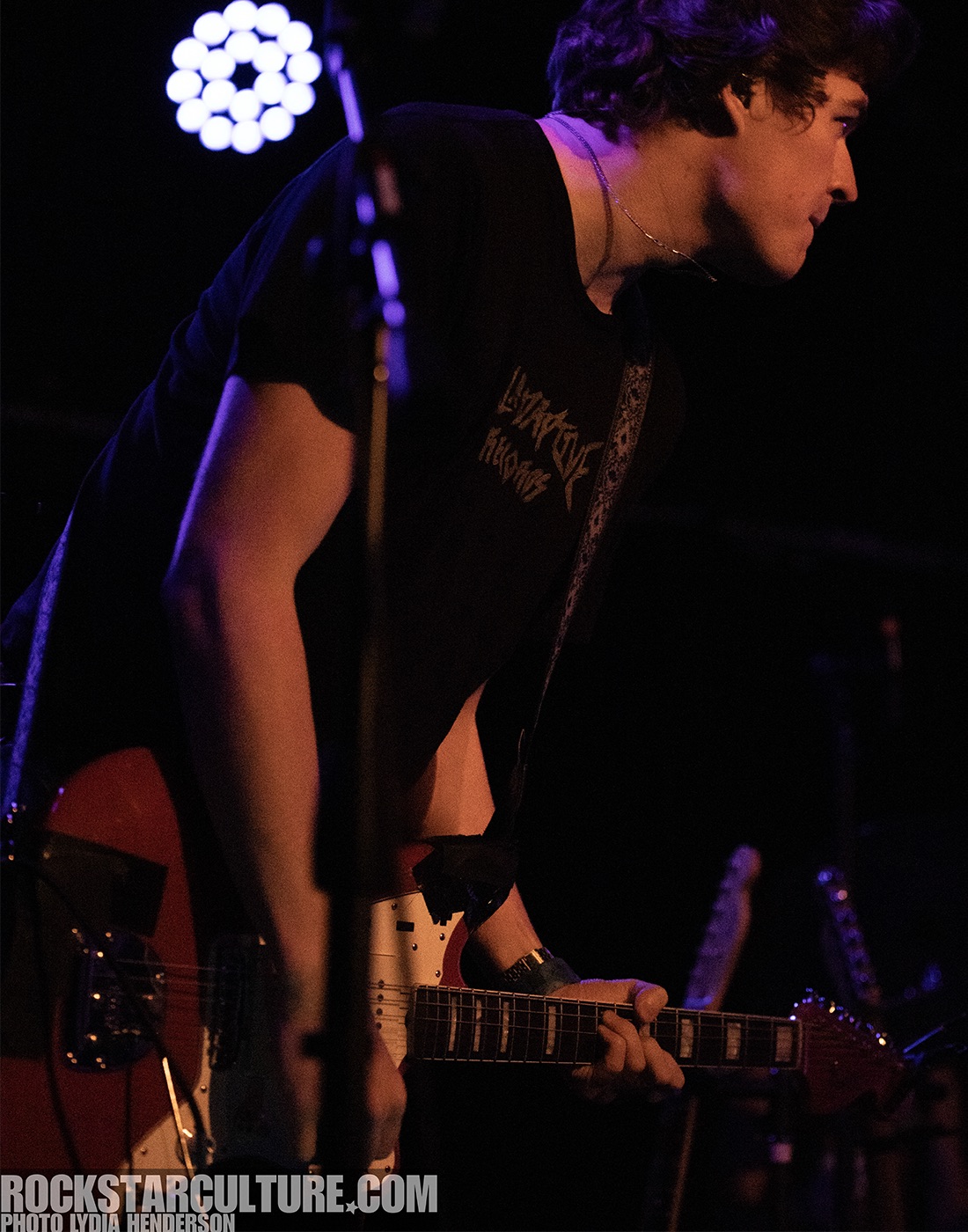 A musician playing an electric guitar on stage, focused and in profile, with a spotlight in the background.
