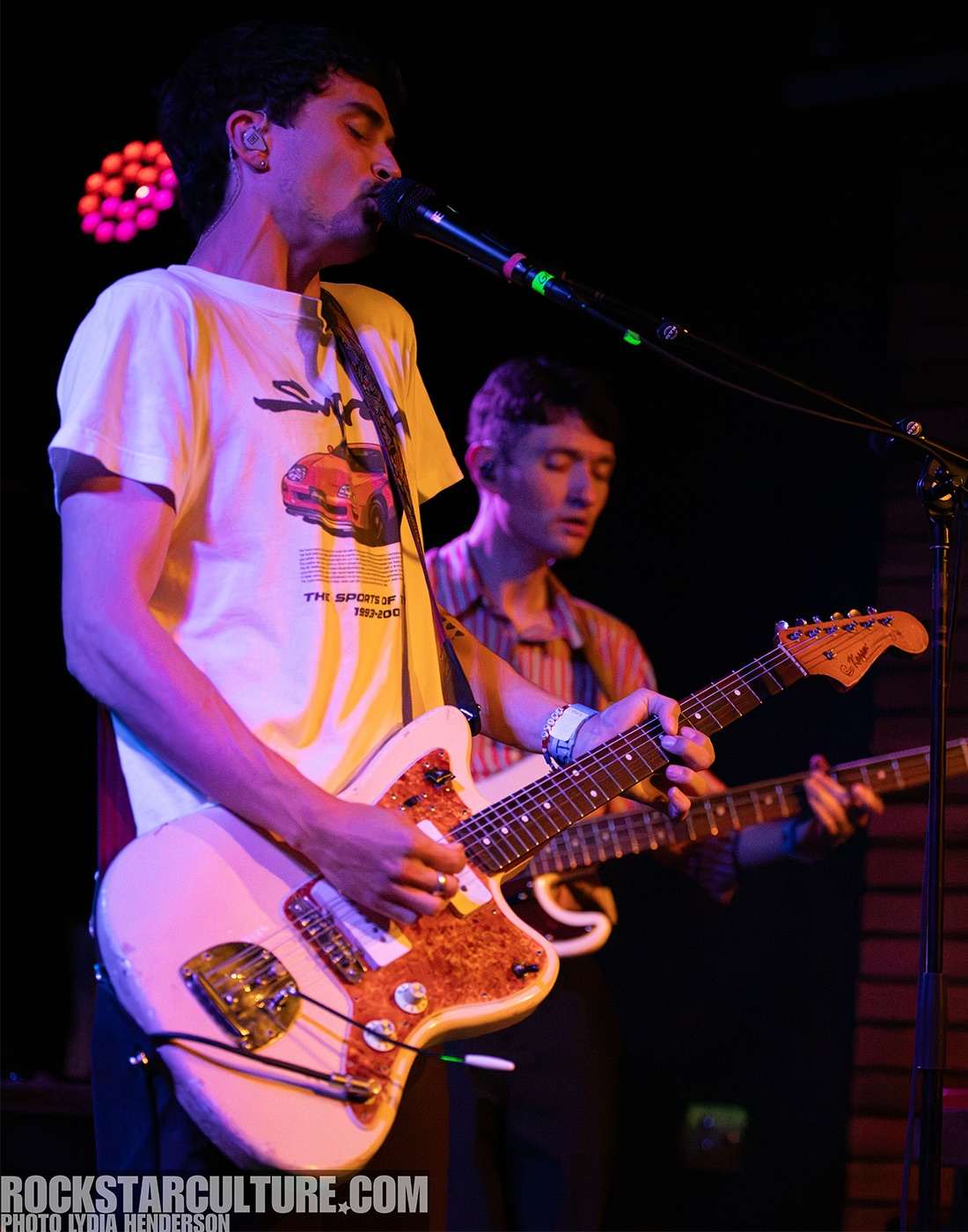 A musician singing into a microphone while playing a guitar on stage, with another band member in the background.