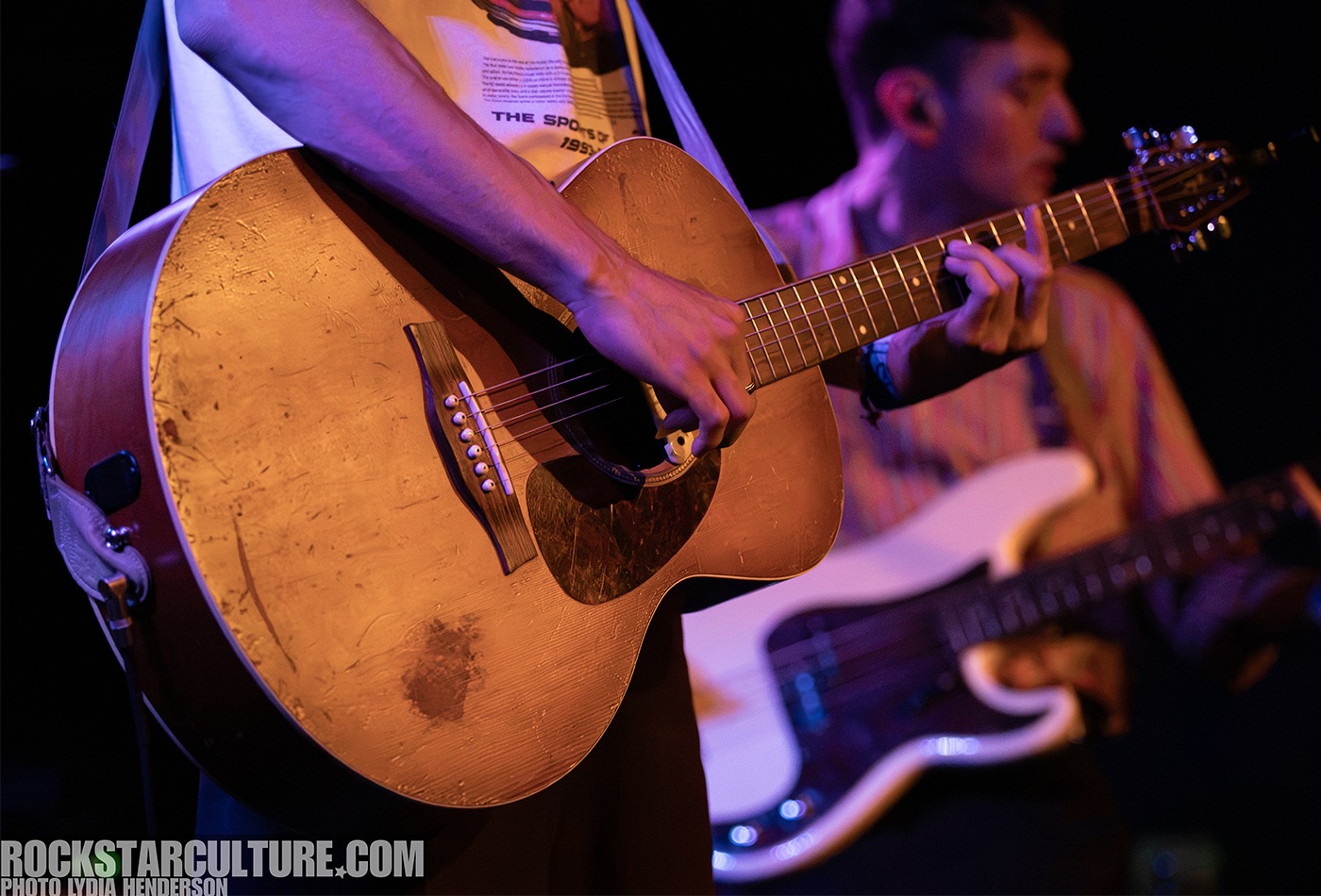 Close-up of a guitarist holding an acoustic guitar while performing on stage, with a bassist partially visible in the background.