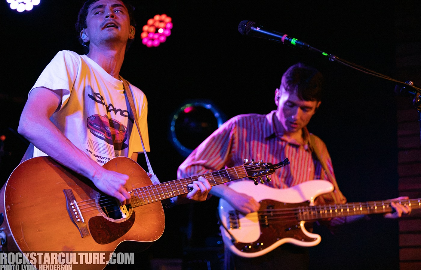 Two musicians performing on stage; one playing an acoustic guitar and the other playing an electric bass guitar.