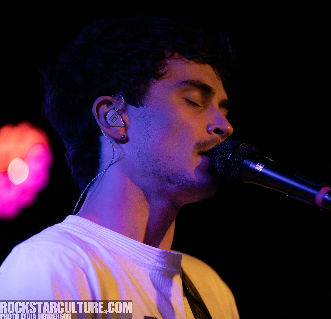 Close-up of a male musician singing into a microphone, with eyes closed and wearing in-ear monitors, illuminated by colorful stage lights.