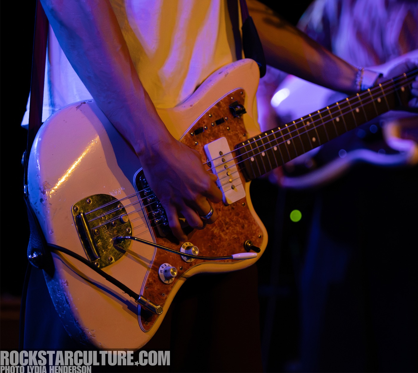 Close-up of a musician's hand playing a vintage-style electric guitar on stage, with colorful lighting in the background.