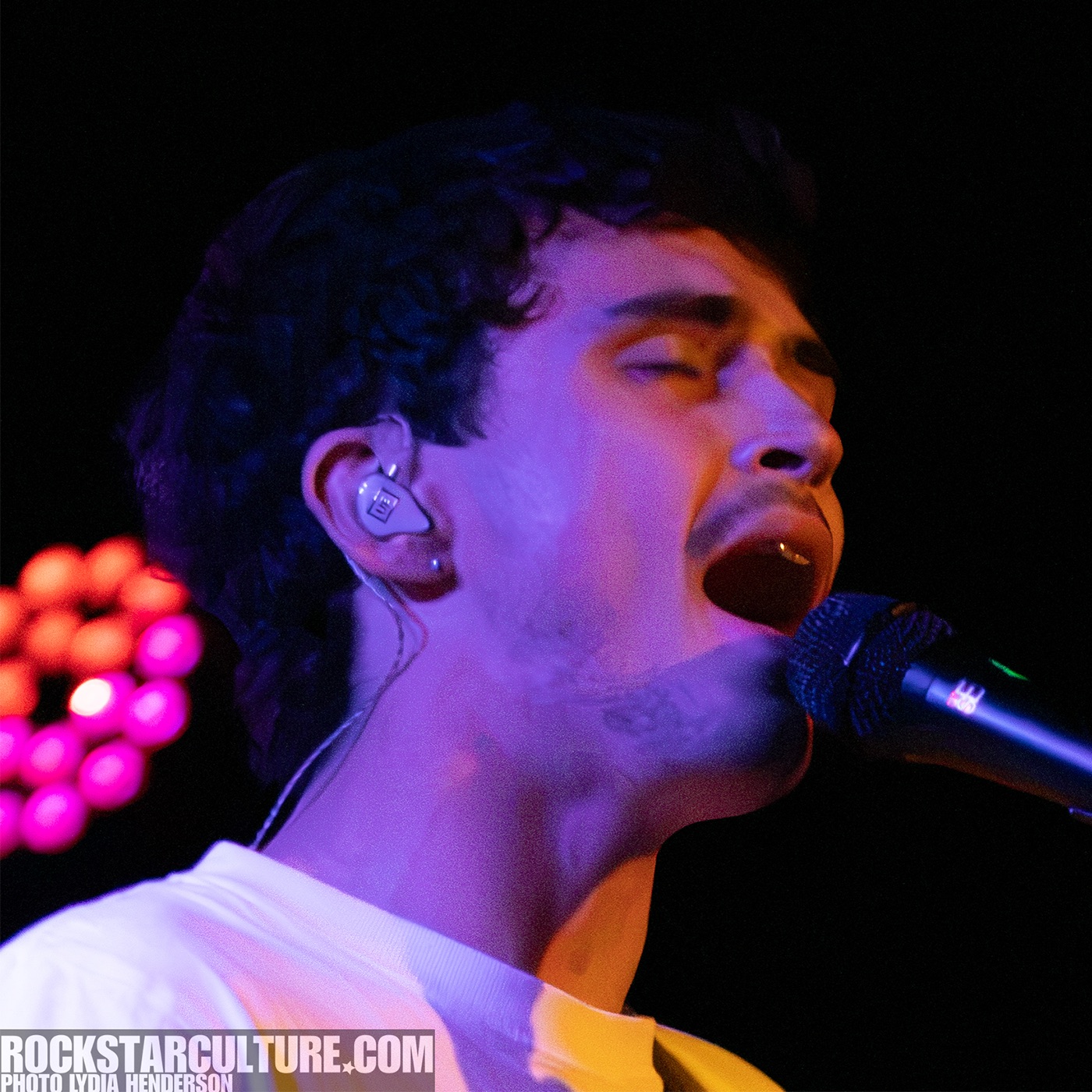 A musician passionately singing into a microphone while wearing in-ear monitors, with colorful stage lighting in the background.