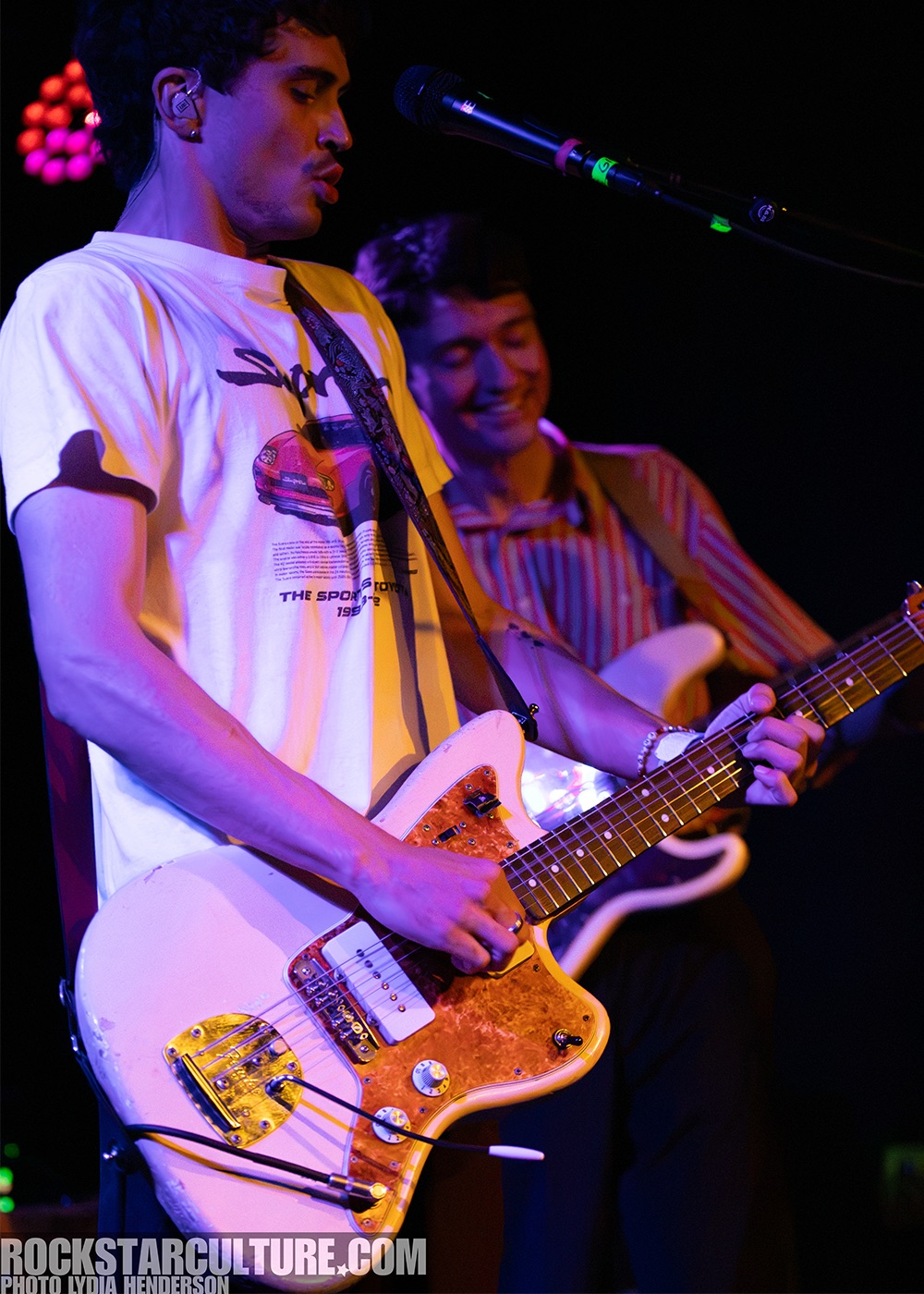 A duo performing on stage, with one guitarist playing a white electric guitar and another musician smiling in the background. The scene is illuminated with colorful stage lights, creating a vibrant atmosphere.