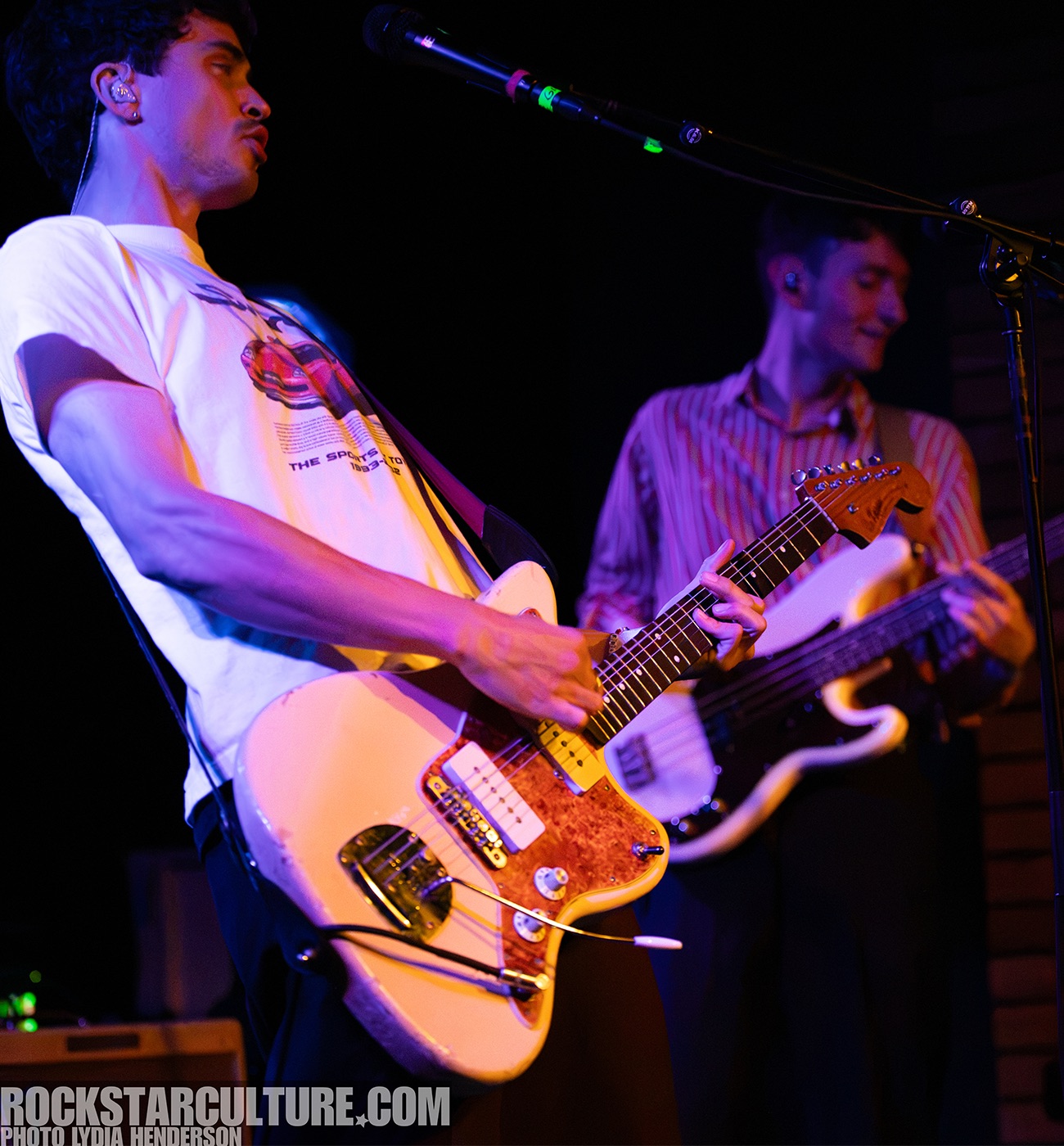 A musician playing an electric guitar on stage, wearing a graphic t-shirt. Another band member is visible in the background, playing a bass guitar.